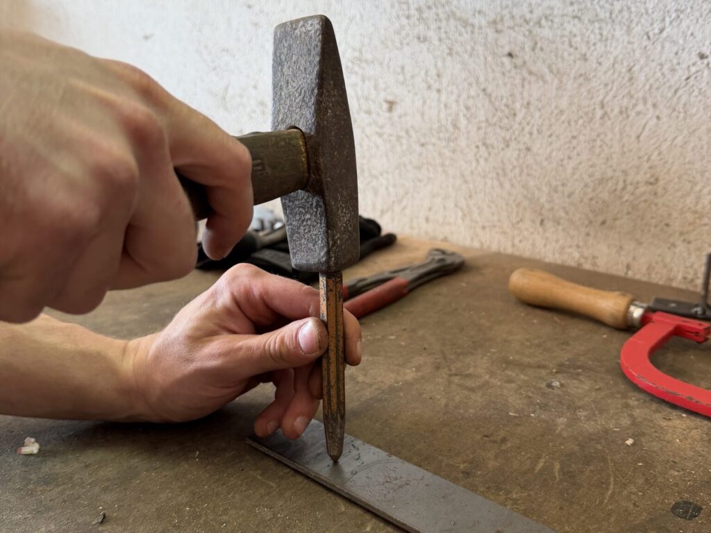 Hände setzen mit Körner und Schlosserhammer einen Ankörnpunkt auf einen Metallstreifen