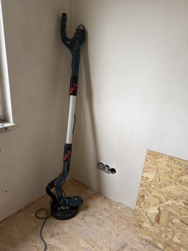 Langhals-Trockenbauschleifer mit Absaugschlauch vor einer frisch gespachtelten Wand, OSB-Platten auf dem Boden