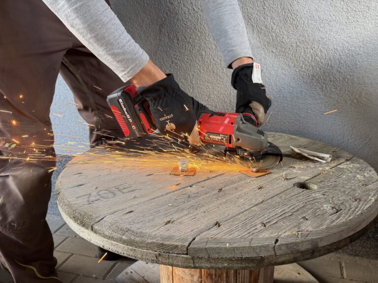 Einhell AXXIO 18/125 trennt einen Metallstift an einer alten Holzkabeltrommel; seitlich fliegen Funken.