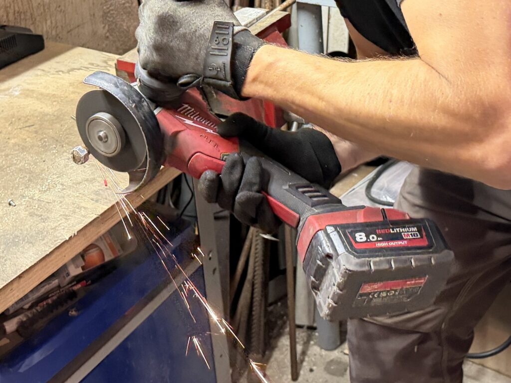 Winkelschleifer mit Trennscheibe an einer eingespannten Metallstange mit Mutter; Funken fliegen nach unten.