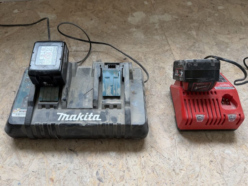 Makita-Doppelladegerät mit 5,0-Ah-Akku und Milwaukee-Ladegerät mit M18-Akku nebeneinander auf einer staubigen OSB-Platte.